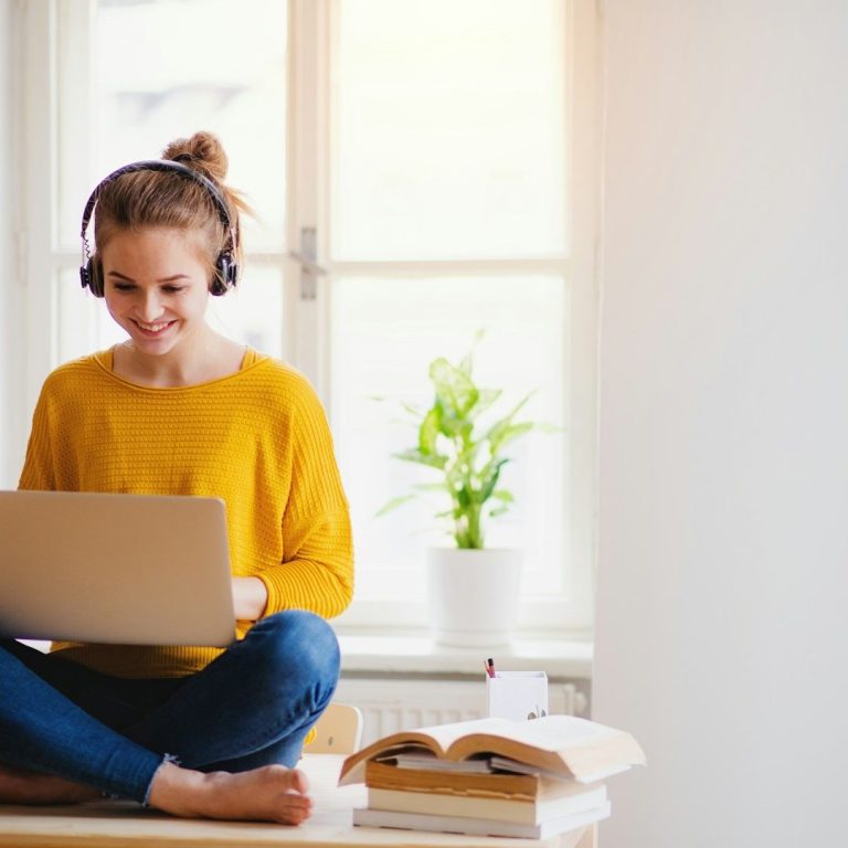 Mujer joven con auriculares, usando una laptop, sentada en una mesa junto a una planta.
