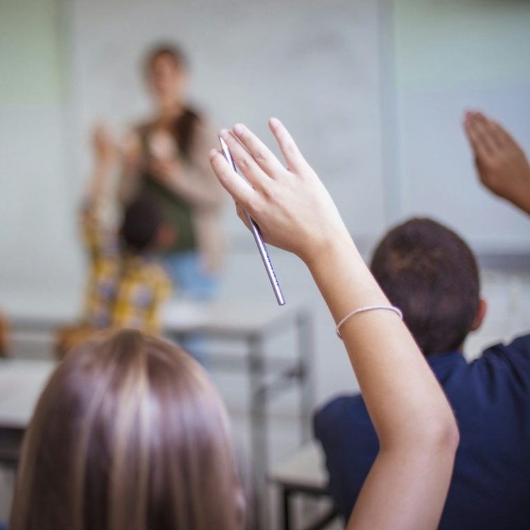 Manos levantadas en un aula mientras una profesora interactúa con los estudiantes.