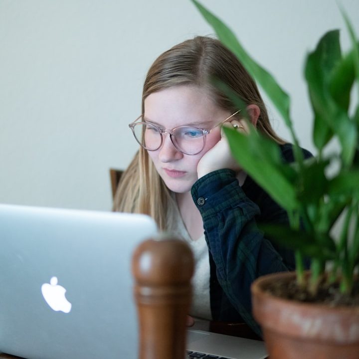 Joven con gafas trabajando en una laptop, apoyando la cabeza en la mano, junto a una planta.