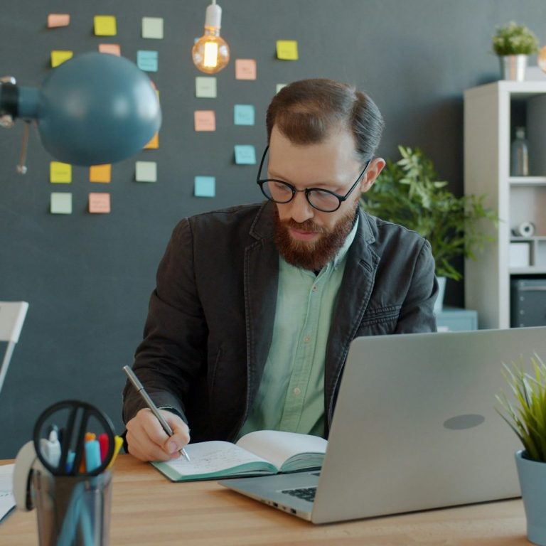 Hombre con barba, gafas y chaqueta, escribiendo en un cuaderno frente a una computadora.